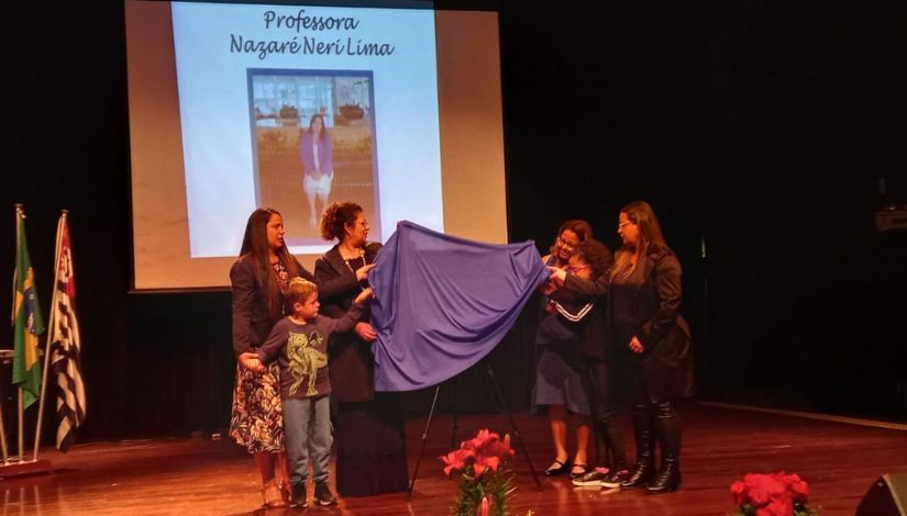 Fotografia do palco com seis pessoas, descerrando a placa de entronização da patronesse e ao fundo no telão projetado a imagem da Professora Nazaré Neri Lima.