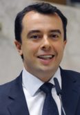 Secretario Bruno Caetano em foto 3x4 em fundo claro, ele veste um paleto azul, camisa branca e gravata azul, ele esta sorrindo.