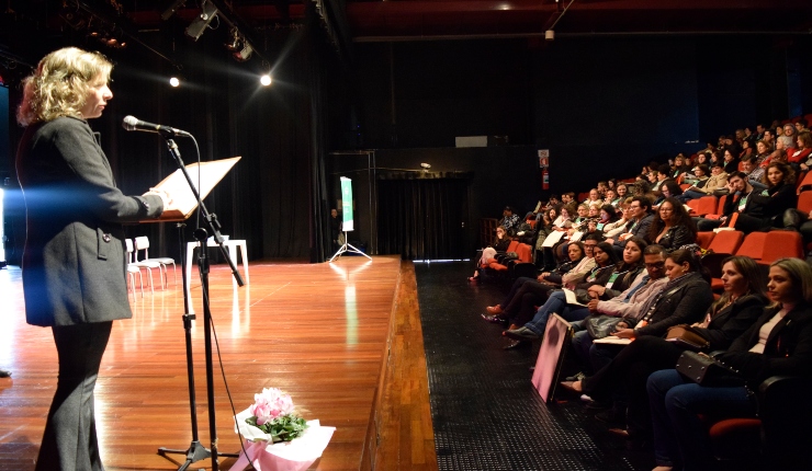 no lado esquerdo da foto no palco uma mulher loira com roupa preta, falando ao microfone. No lado direito da imagem, publico do auditório.