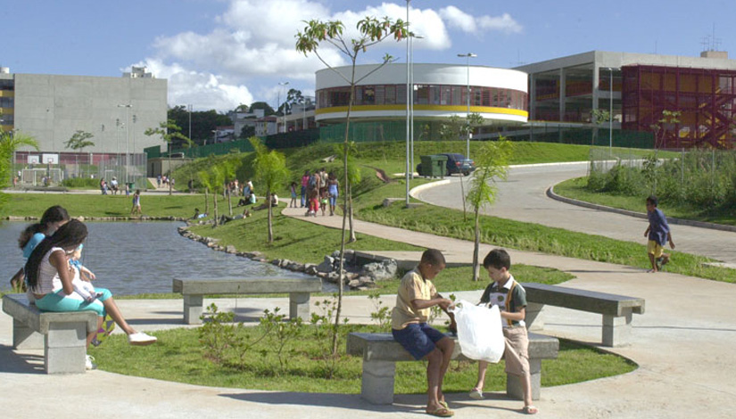 Foto do CEU Butantã. Em primeiro plano, crianças sentadas em bancos de concreto. Ao meio, à esquerda um lado, ao centro um caminho com pessoas caminhado, à direita uma rua. Ao fundo da foto, os prédios do CEU, os das pontas são retangulares e o do centro é circular
