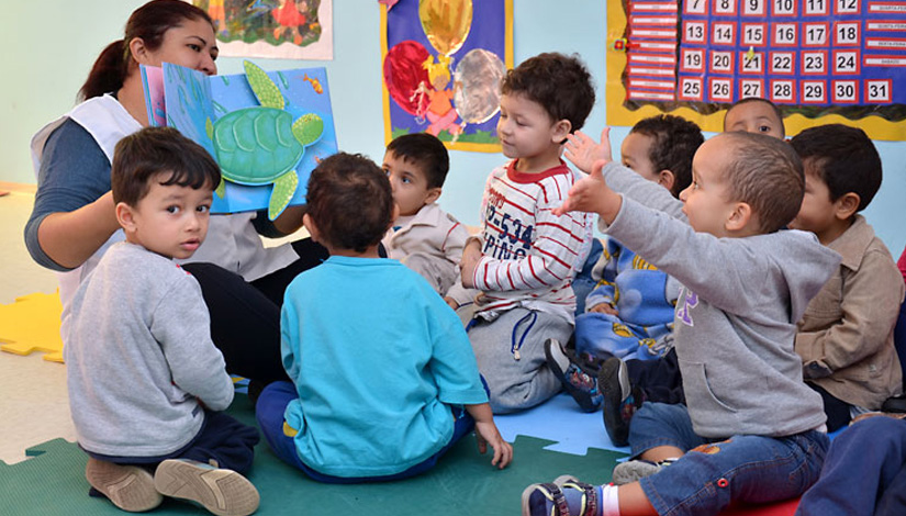professora sentada no chão, mostra um livro para as crianças também sentadas no chão fazendo um círculo. 