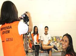 No primeiro plano, uma garota de costas, com o colete laranja do projeto imprensa jovem fazendo filmagens, do outro lado, uma garota olha sorridente pra foto. No segundo plano um estudante entrevistando seu colega
