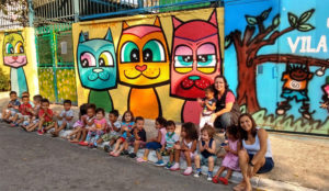 Crianças do CEI sentadas na calçada em frente ao muro pintado com três gatos coloridos.
