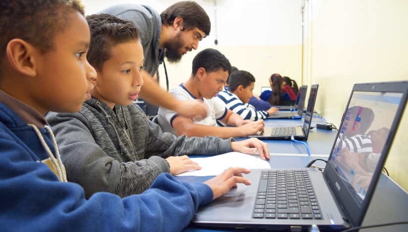 Fotografia mostra crianças em um laboratório de informática usando notebooks. Um professor orienta um estudante que está sentado à mesa