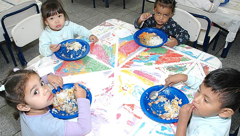 Quatro crianças comendo a merenda em uma mesa quadrada.