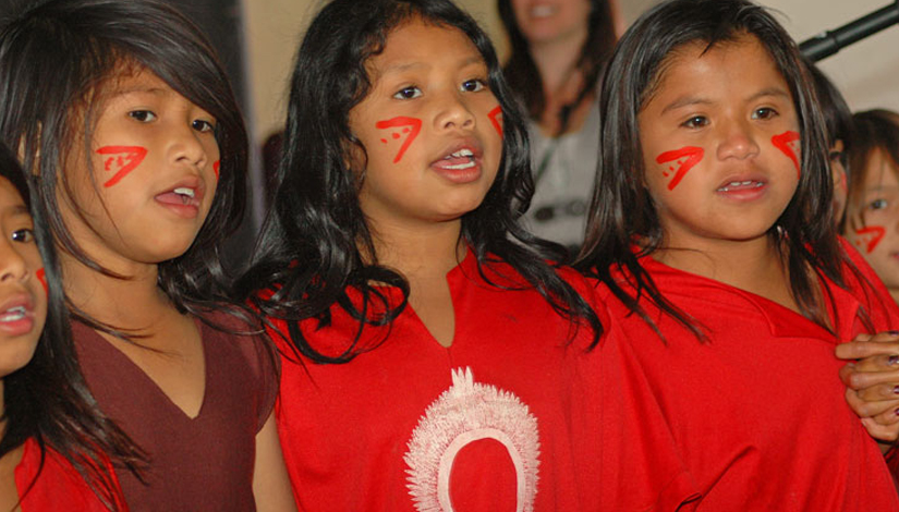 Três meninas guarani em fila, de camisa vermelha, cantando.