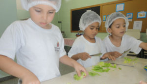 Três meninos cortando folhas de alface, atividade educacional proposta pela escola para conscientização das crianças sobre alimentação.