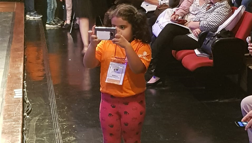 Imagem de uma criança de pé em frente a platéia tirando foto com um celular.