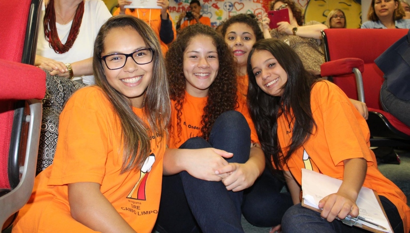 Uma fotografia mostra 4 garotas com camiseta laranja Imprensa Jovem juntas sorrindo para foto.