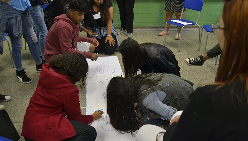 Imagem mostra alunos desenhando uma silhueta de uma pessoa em um papel no chão.