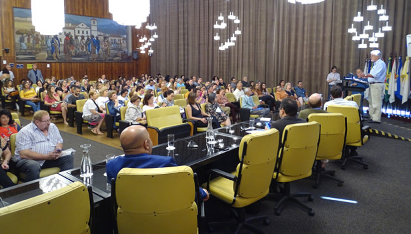 Imagem mostra várias pessoas sentadas em cadeiras amarelas participando do seminário sobre a FUNDEB.