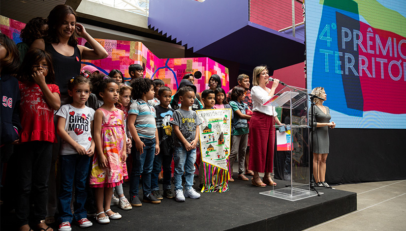 Imagem com crianças sobre o palco e uma mulher com microfone na mão ao lado delas.