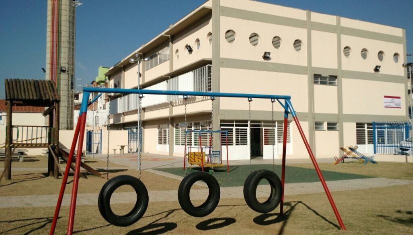 Fotografia de uma escola municipal. prédio com três pavimentos e um parque infantil em primeiro plano