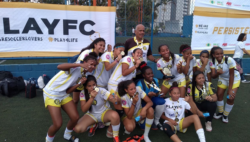 Imagem das meninas do futebol feminino posando para foto com as medalhas e o troféu.