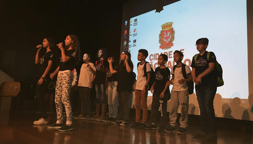 Imagem de alunos sobre o palco, e atrás deles um telão.