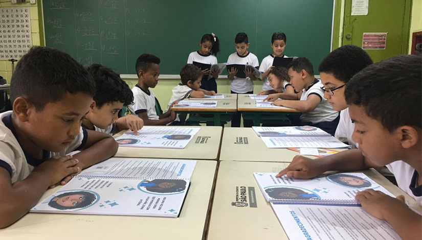 Imagem de uma sala de aula com alunos sentados lendo o livro, e de fundo alunos de pé com livros em suas mãos.