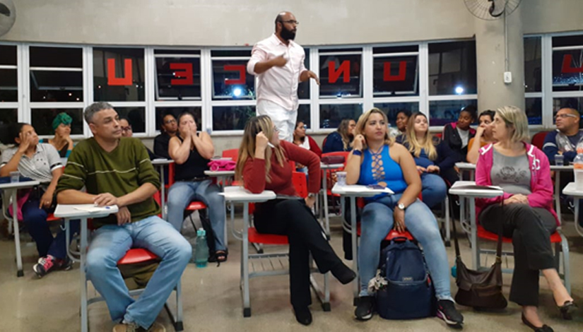 Imagem de uma sala de aula com pessoas sentadas e um homem no meio de pé.