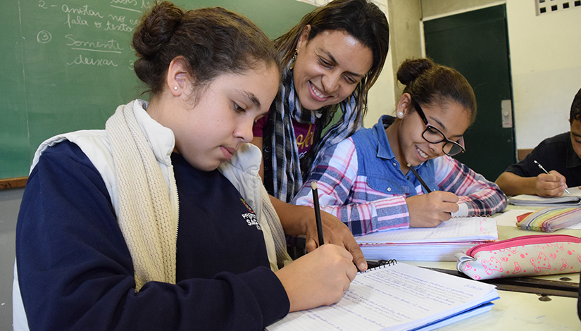 Imagem com 2 alunas escrevendo em um caderno e a professora dando instruções ao lado das duas, ao fundo uma lousa de sala de aula.