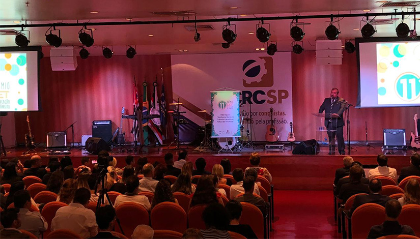 Imagem tirada de trás da platéia presente no evento de Premiação por Projetos de Educação no Trânsito, e no fundo da imagem o palco.