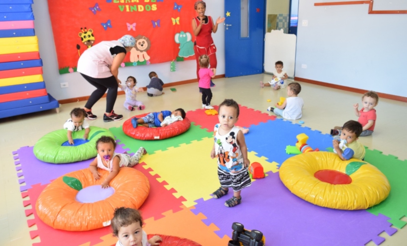 foto de uma sala com bebês, o chão é forrado com placas coloridas de emborrachado e alguns almofadões. Alguns bebês olham para a câmera. Ao fundo, Duas mulheres interagindo com eles