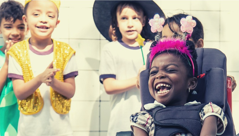 Imagem com alunos aplaudindo ao fundo e na frente uma criança sorrindo com uma tiara rosa.
