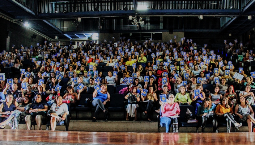 Imagem mostra mais de 100 educadores sentados no teatro da DRE Guaianases levantando um fôlder utilizado na formação do evento.