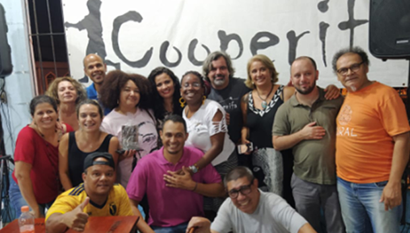 Imagem de um grupo de pessoas posando para a foto, e ao fundo um cartaz escrito "Cooperifa".
