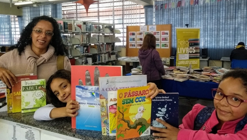 Imagem mostra duas garotas sorrindo segurando livros ao lado de uma educadora na Biblioteca do CEU Inácio Monteiro