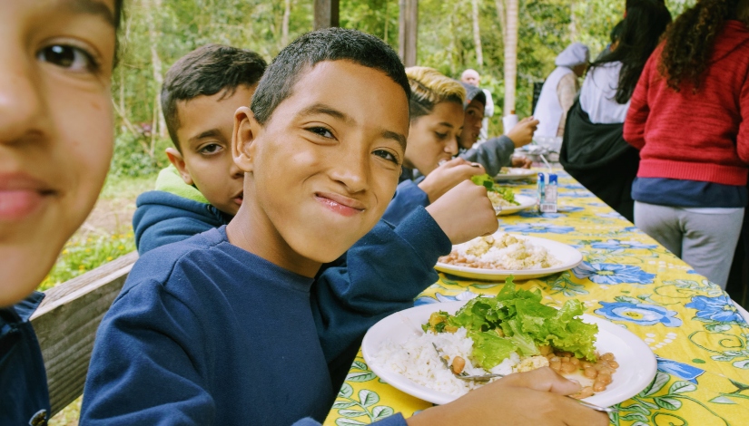 Imagem mostra aproximadamente cinco estudantes sentados na mesma mesa, um ao lado da outro se alimentado, um deles, um menino, está posando para foto e comendo.