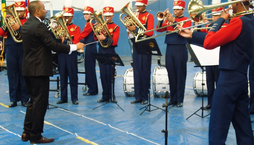 Imagem mostra oito estudantes realizando um apresentação musical tocando instrumentos de metais sendo conduzidos por um Professor de música que está à esquerda da imagem.