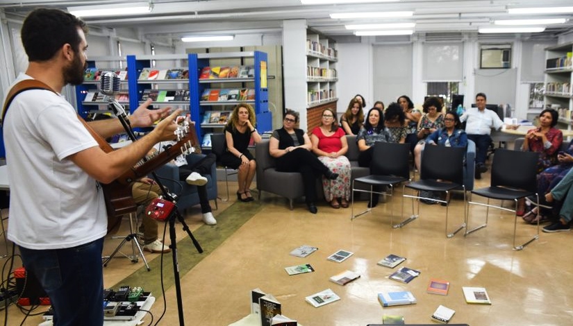 Imagem mostra uma pessoa em pé realizando uma apresentação musical, ao lado direito, um grupo de aproximadamente 20 pessoas acompanham sentados a apresentação, no chão, alguns exemplares de livros.