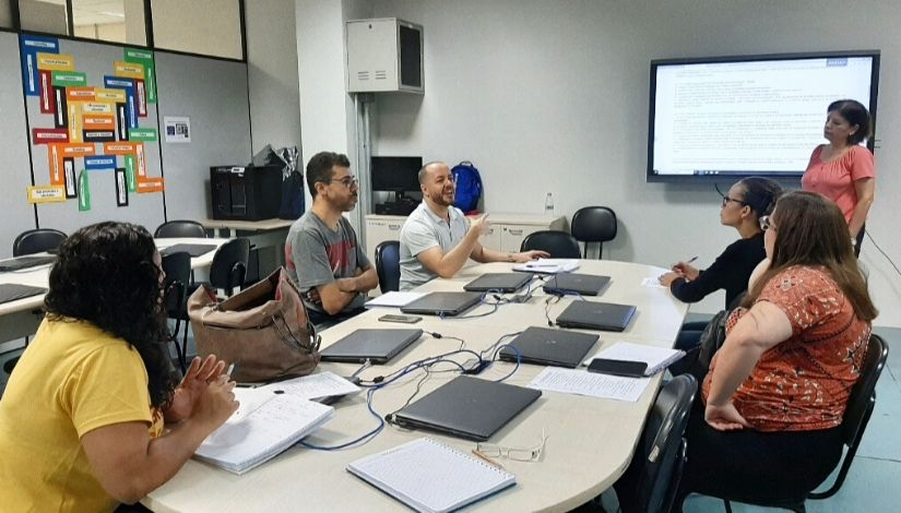 fotos de pessoas reunidas em volta de uma mesa. Há uma tela projetada na parede e laptops em cima da mesa