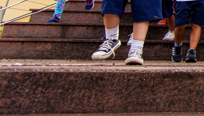 Fotografia dos pés dos alunos descendo uma escada. Alunos usam bermuda do uniforme e tênis.