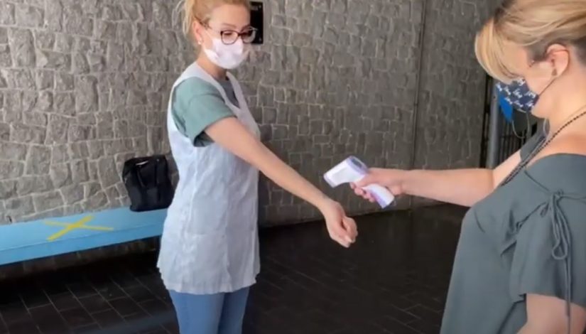 Fotografia com uma mulher de avental e máscara tendo a temperatura aferida com termômetro digital no punho por outra mulher na portaria da escola