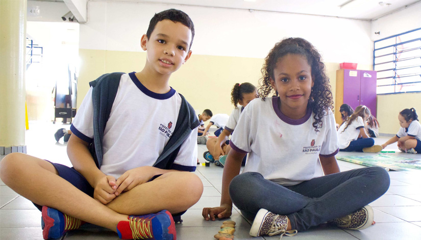 Dois alunos da rede, um menino e uma menina sentados no chão do pátio usando uniformes.