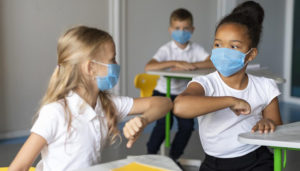 Duas meninas na sala de aula usando máscara de proteção e uniforme se cuprimentando com os cotovelos.