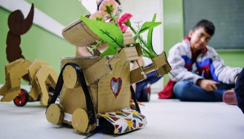 robô motorizado feito com materiais recicláveis e criança ao fundo segurando o controle remoto
