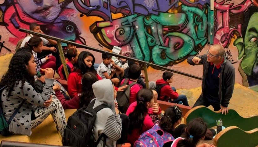 Imagem de um grupo de estudantes com o professor em uma aula de Geografia pelo bairro. Estudantes sentados em uma escadaria, professor à frente conversando com eles, ao fundo, parede com grafites muito coloridos.