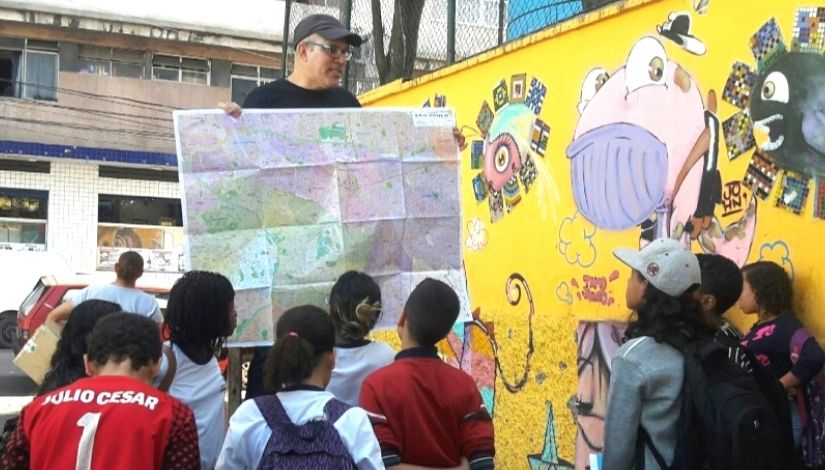 Imagem mostra professor de Geografia dando aula em uma calçada nas ruas de SP. Ele está segurando um mapa aberto em sua frente. Um grupo de estudantes observam o mapa e olham para o professor. Ao fundo um muro amarelo com desenhos de grafite.