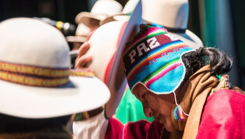 fotografia de homem com trajes andinos em apresentação. Ele faz gesto com chapéu enquanto usa uma touca com a palavra paz