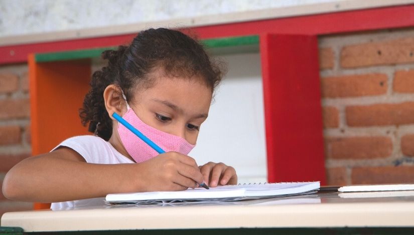 Estudante escrevendo em seu caderno com lápis grafite. Ela usa máscara de proteção individual.