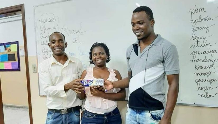 Professor e estudantes em frente a lousa com palavras escritas em Crioulo. Eles seguram uma caixa de chocolate.