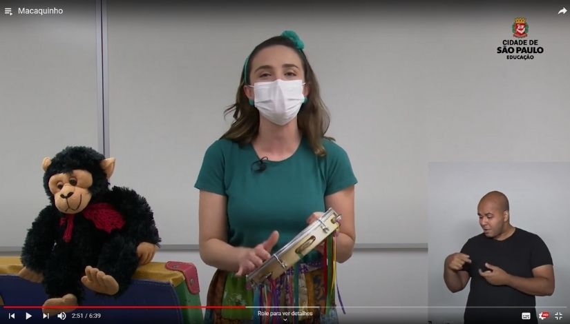 imagem de vídeo de professora fazendo contação de história utilizando pandeiro e macaco. 