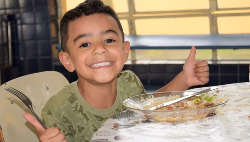 Menino em momento de alimentação na escola. Ele está sorrindo, sentado à mesa e faz sinal de joinha com as duas mãos. Sobre a mesa um prato de vidro com arroz, feijão e salada.