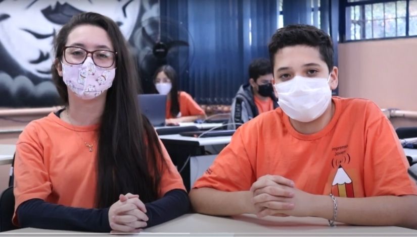 foto de estudantes com máscaras usando camisetas na cor laranja da imprensa jovem sentados lado a lado apresentando telejornal