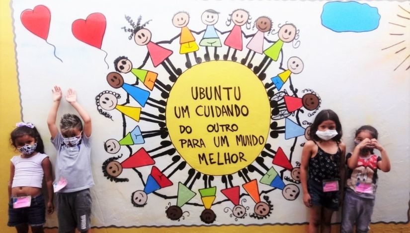 crianças ao lado de um painel pintado na parede com a representação de diversas pessoas em roda em volta da frase 
