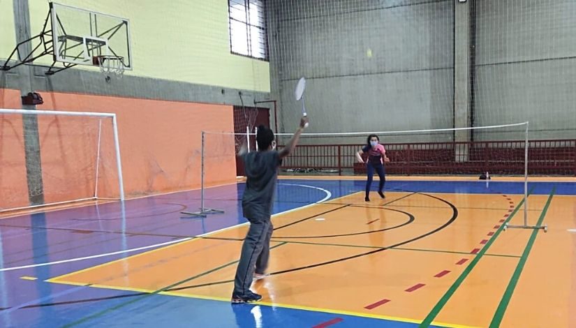 Imagem mostra uma estudantes e um estudante jogando badminton na quadra da escola.