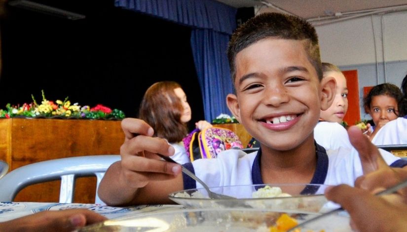 Fotografia de estudante fazendo sua refeição na escola. Ele está sorrindo e usa colher e prato de vidro. Atrás dele, outras crianças estão sentadas à mesa também fazendo suas refeições.