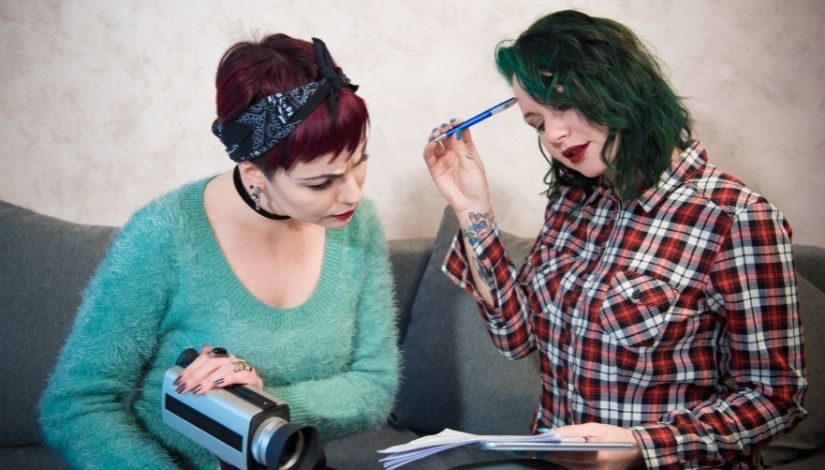 Imagem de duas mulheres: uma de cabelo curto vermelho com lenço amarrado, ela usa blusa de lã verde claro e tem uma câmera nas mãos; a outra tem cabelos médios coloridos de verde, ela veste camisa de manga longa xadrez e está segurando um tablet, folhas de papel e caneta. As duas olham para os papeis que estão sobre o colo de uma delas.