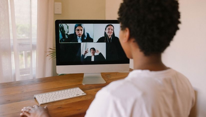 Imagem de uma pessoa de costas, ela está vira para a tela de um computador onde aparecem três mulheres em reunião.